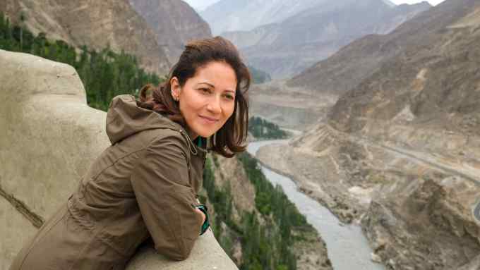 A woman with long brown hair wearing an anorak smiles as she leans over a wall and looks along a wide brown mountainous valley with a river snaking through it