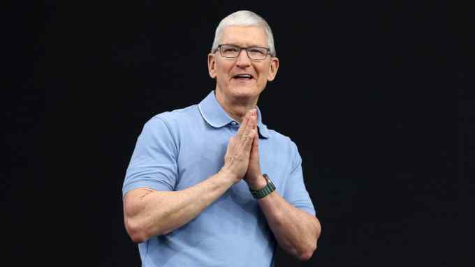 Apple CEO Tim Cook in light blue polo shirt clasps his hands in front of his chest as he address the audience