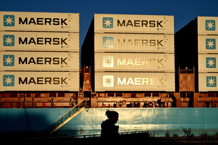 Dramatic evening light illuminates a cargo ship stacked with multiple layers of Maersk shipping containers, with a shadowed figure walking in the foreground