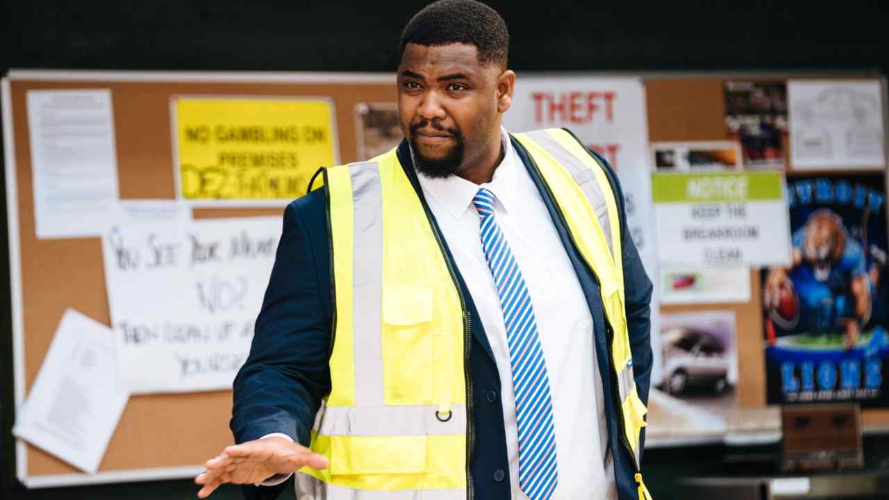 A man wearing a suit and tie and yellow hi-viz jacket holds out one hand warningly as he stands before an office bulletin board