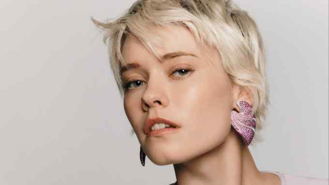 A close-up portrait of a woman with short platinum blonde hair, wearing statement pink gemstone earrings. She is dressed in a lavender top with a keyhole cutout at the chest