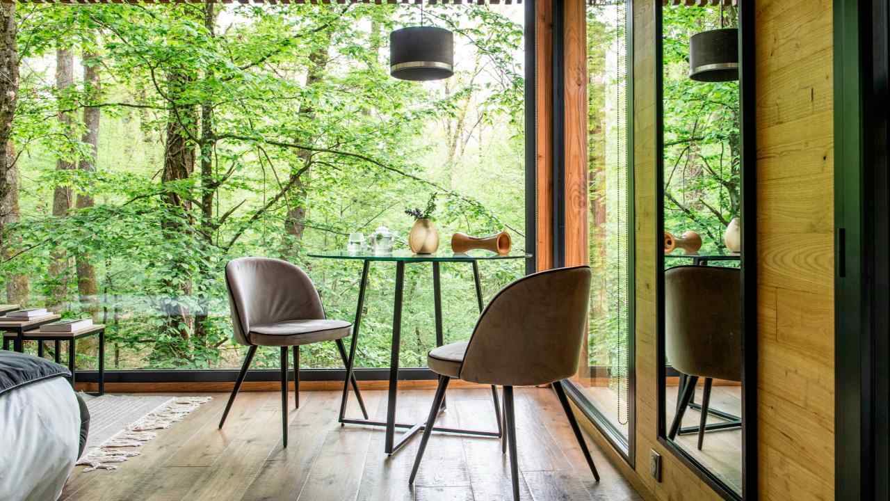 A table and two chairs on a wooden floor, by floor-to-ceiling windows with a view of trees in leaf