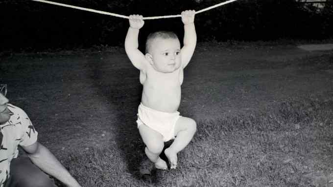 A black and white photo of a baby in a nappy hanging on to a line in a garden