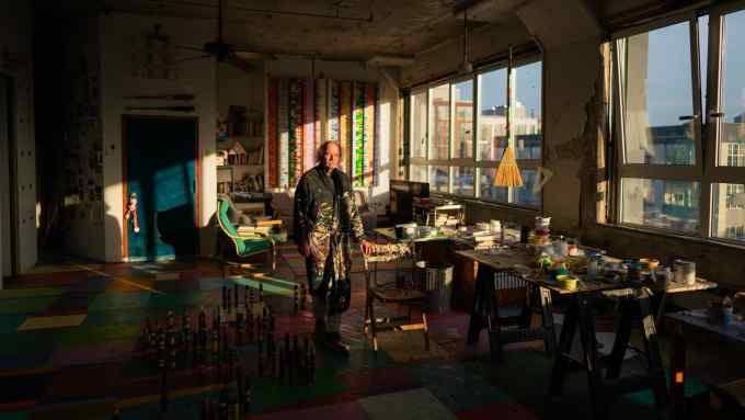 A man in paint-spattered overalls stands in a loft studio as sunlight streams in through the windows