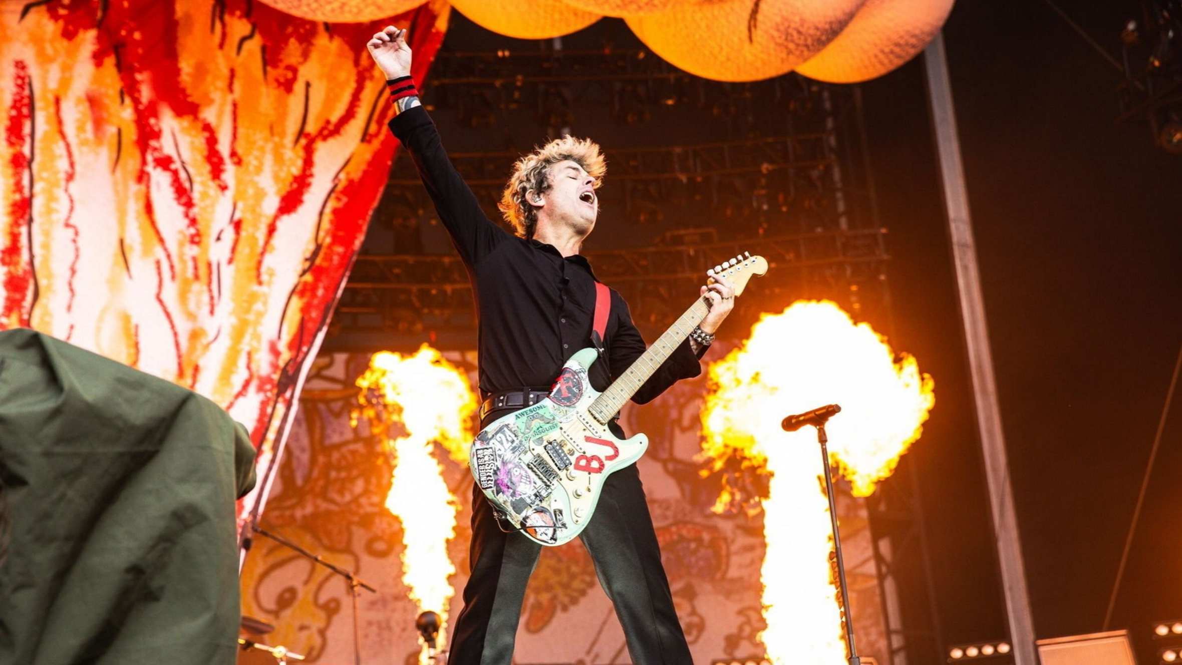A short man with peroxided hair wearing black holds a guitar and raises one arm in triumph as firejets flame behind him