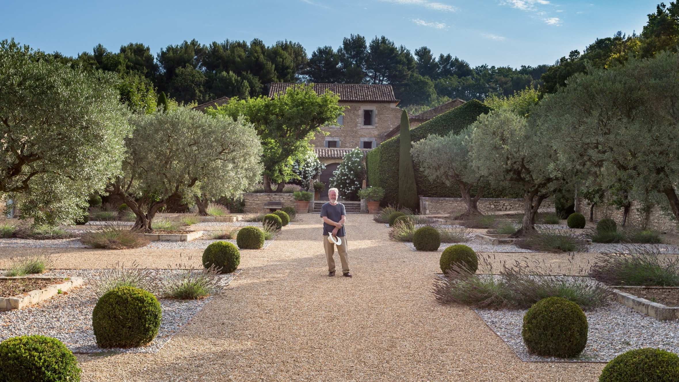 Ridley Scott, with grey hair and beard, holding a hat, stands smiling in the gardens of a large house in the country