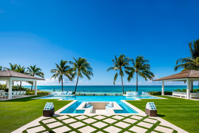A pristine seaside garden with a central swimming pool surrounded by lush green grass and patterned stone paths, set against the backdrop of a turquoise sea and clear sky