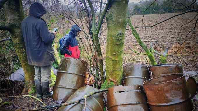 Two people in raincoats with the hoods up stand by rusty barrels