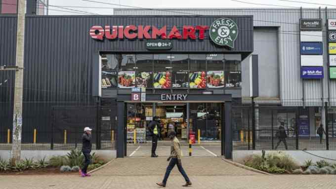 The exterior of a Quick Mart supermarket is shown, with the store’s name in bold red letters and a “Fresh & Easy” logo next to it. The sign above the entrance indicates the store is open 24/7. Large windows display promotional posters featuring fresh produce and other food items. A few people are seen walking outside, and a security guard stands at the entrance