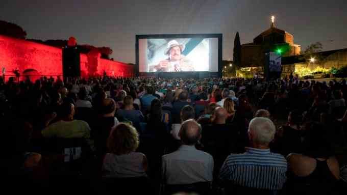 Crowds watching the giant screen of open-air cinema at Parco degli Acquedotti after dark, with ancient walls illuminated red to the left of the image