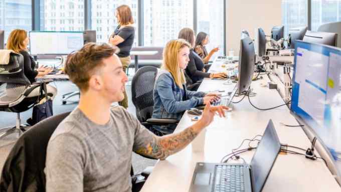 A busy office setting with multiple employees at desks using computers. The foreground shows a young man with short hair and a tattooed arm, while the background features various colleagues, including a woman in a denim jacket