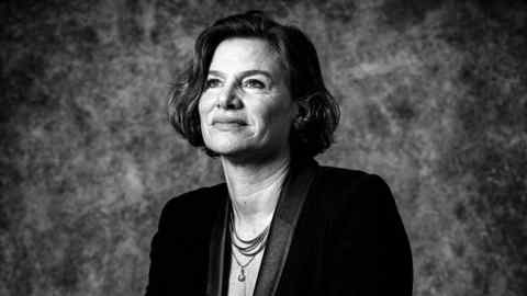 Black and white portrait of Mariana Mazzucato wearing a long, patterned skirt and black jacket sitting on a stool with her legs crossed
