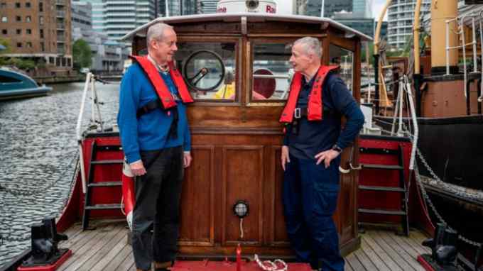 Two elderly men wearing life jackets stand on the deck of a vintage boat docked in a modern city marina, surrounded by tall buildings and other boats