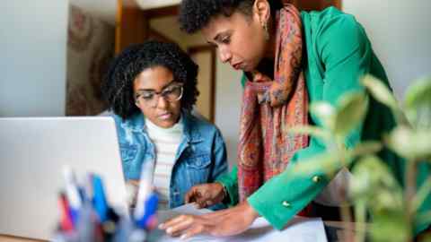 Two businesswomen using a laptop and discussing paperwork while working together at a desk in their home office