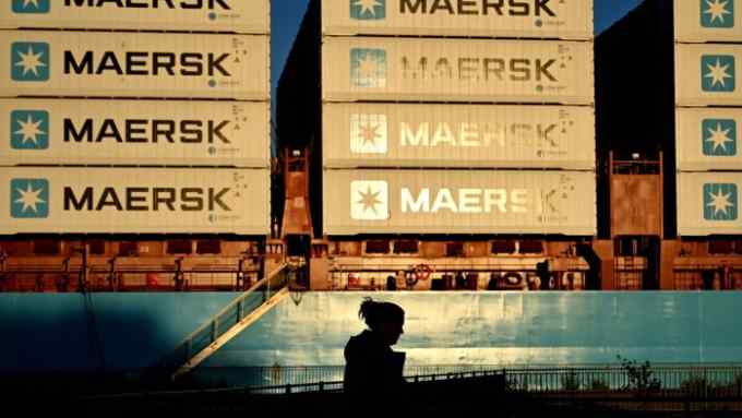 Dramatic evening light illuminates a cargo ship stacked with multiple layers of Maersk shipping containers, with a shadowed figure walking in the foreground