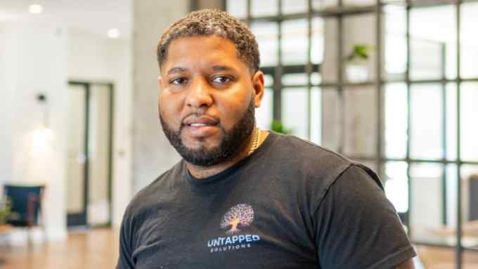 Portrait of a Black man with short black hair, standing in an office environment. He wears a black T-shirt with a logo that reads ‘Untapped Solutions’ and is tattooed on both arms
