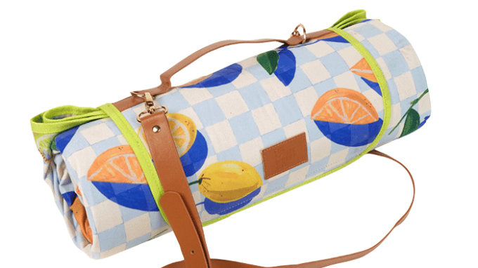 A rolled-up picnic blanket with a blue and white checkered pattern featuring colorful fruit designs, secured with brown leather straps and a carrying handle