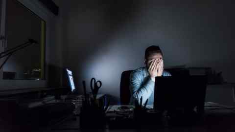 An exhausted male office worker sitting at his desk in a dark room, face covered by his hands, with visible stress, beside a computer showing business data