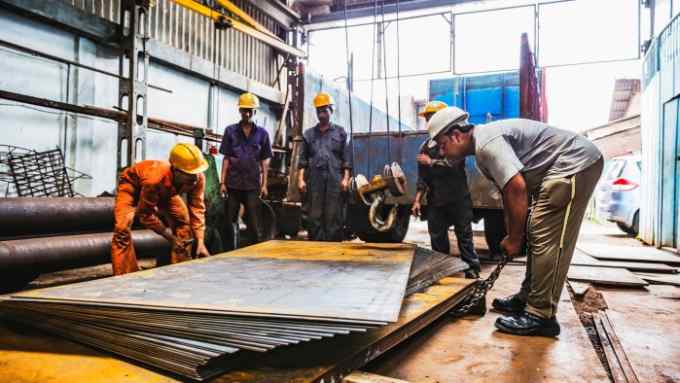 A group of workers in a metal workshop are handling large sheets of metal. They are wearing protective gear, including helmets and overalls. One worker in an orange jumpsuit is bending down to lift a sheet, while another in a gray shirt is using a tool to assist