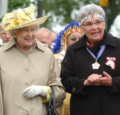 Wearing a beige rain jacket, The Queen walks beside an Indigenous woman wearing a beaded headband and two medals. 