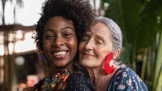 Two women embracing amicably, pressing their heads together, one behind the other, both smiling.