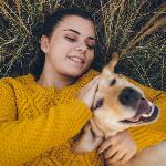 A woman in a bright yellow sweater laying in some tall grass outside, hugging her dog, a golden lab, and petting his head.