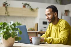 A man in a yellow shirt sits in a kitchen at a table with a plant, a laptop, and a mug