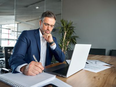 A man in a suit writing in a notebook in front of a laptop in an office. 