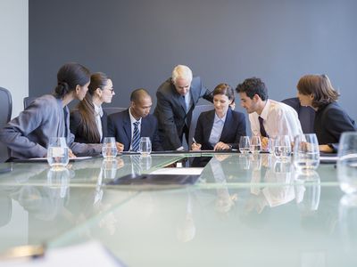 Businesspeople gathered around a glass conference table watch as one person signs a business agreement.