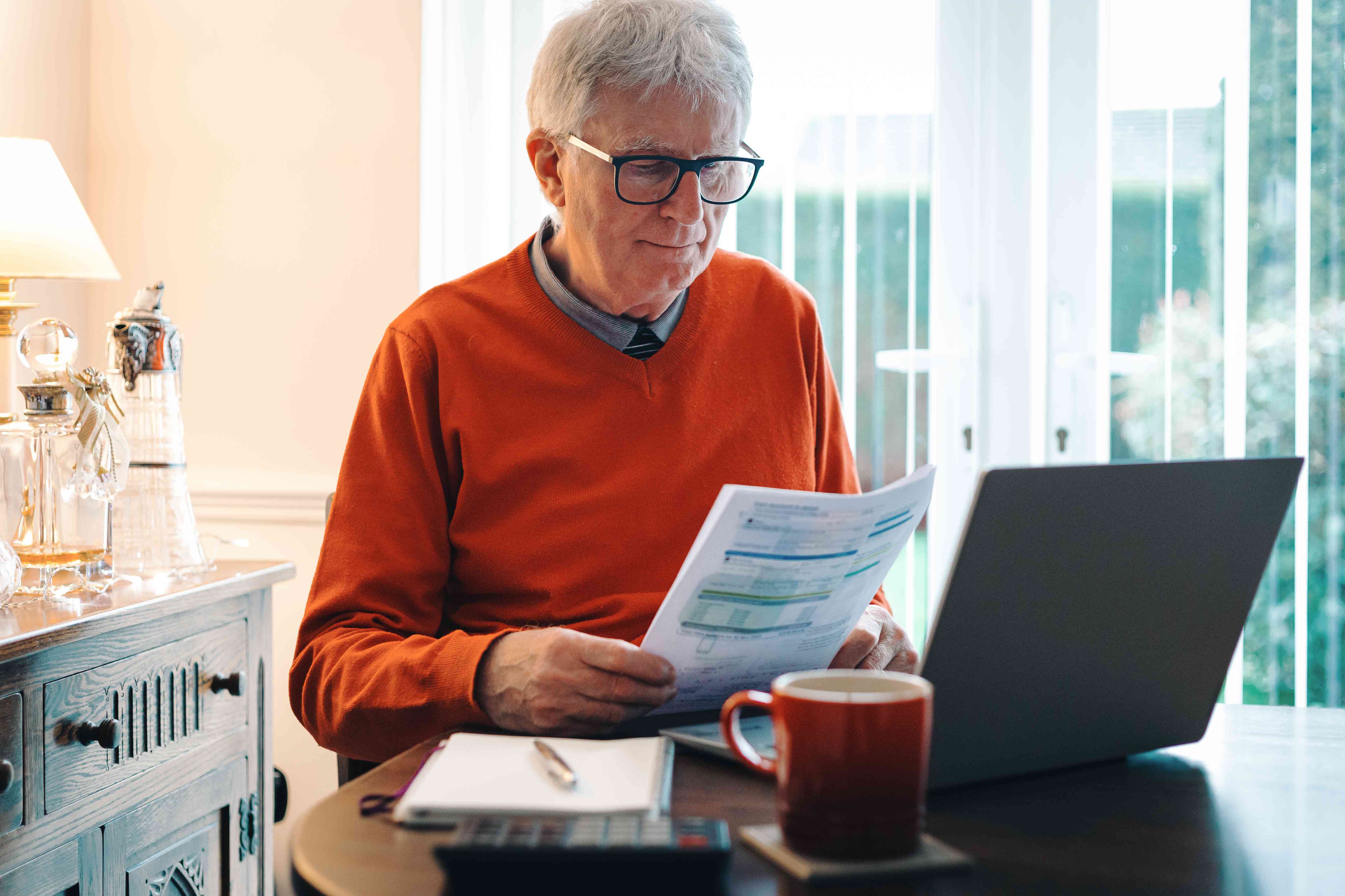 A retiree sitting at home in front of a laptop and reading over investment portfolio paperwork.