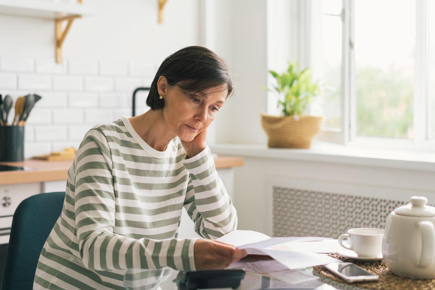 Woman at a kitchen table reading mail in front of a teapot next to an open window. 