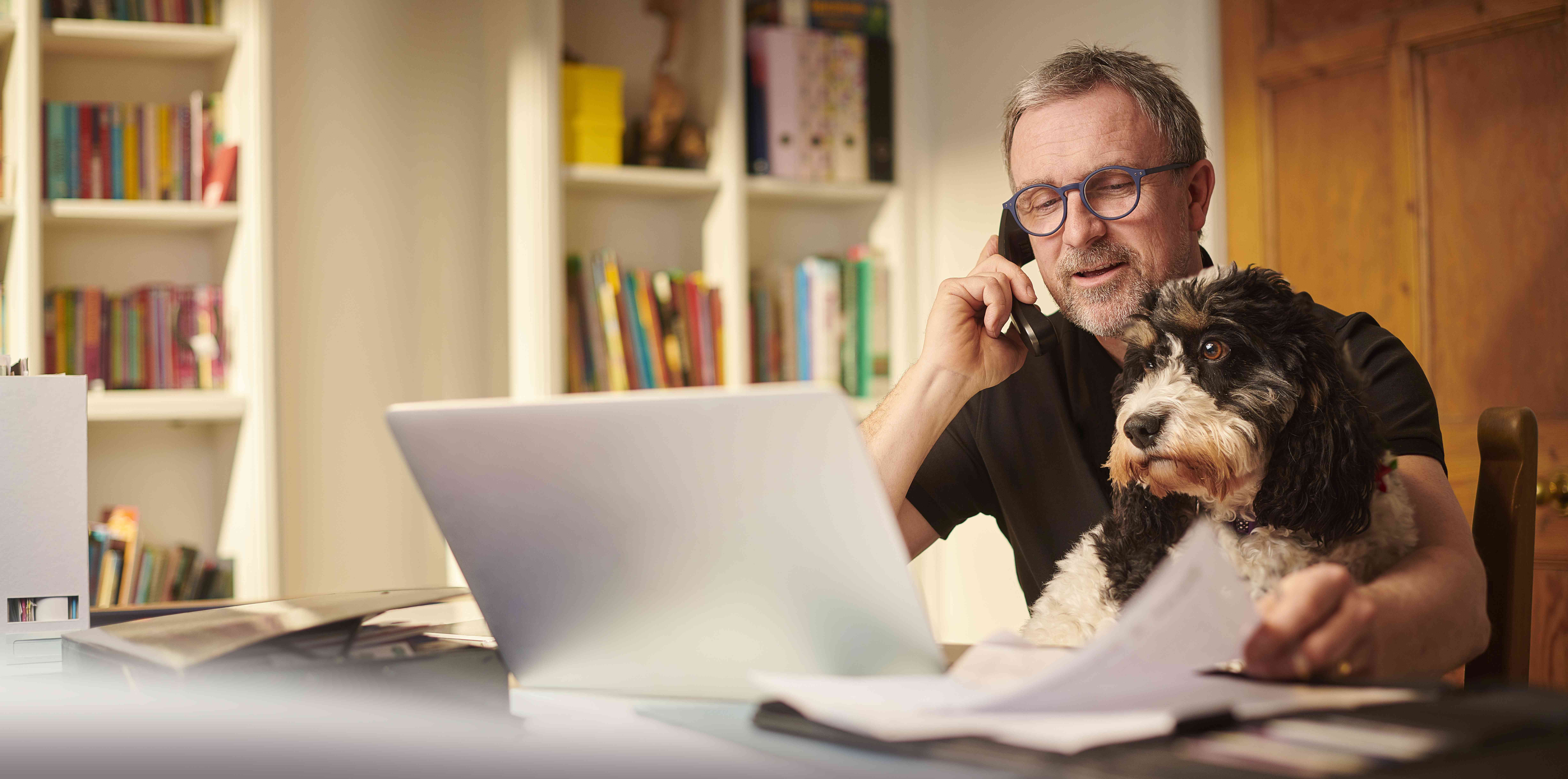 A man works from home. He's talking on the phone, and looking at a laptop screen while a fluffy mid-sized dog sits on his lap..