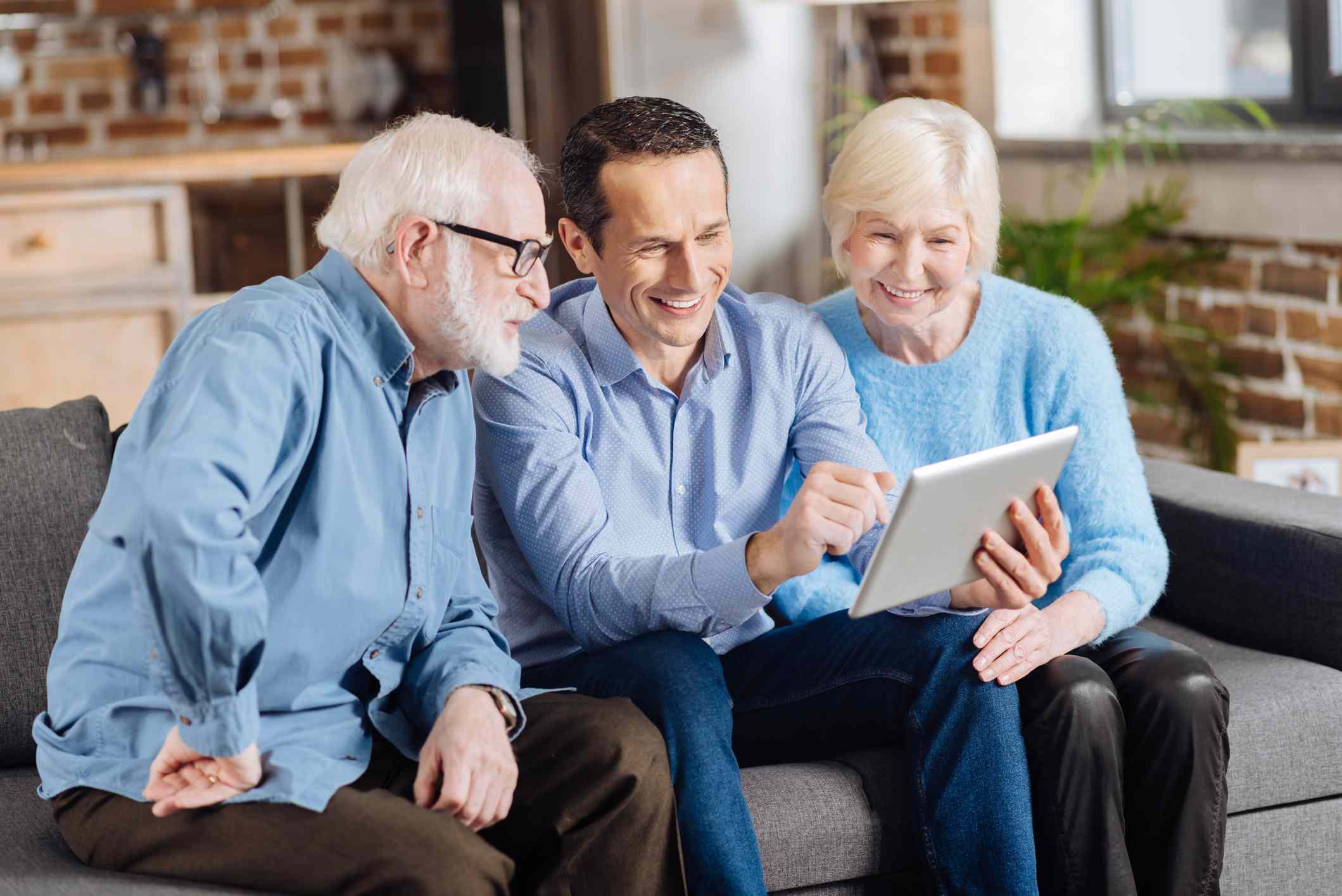 A young man sitting on the couch between his parents and teaching them how to use a tablet.