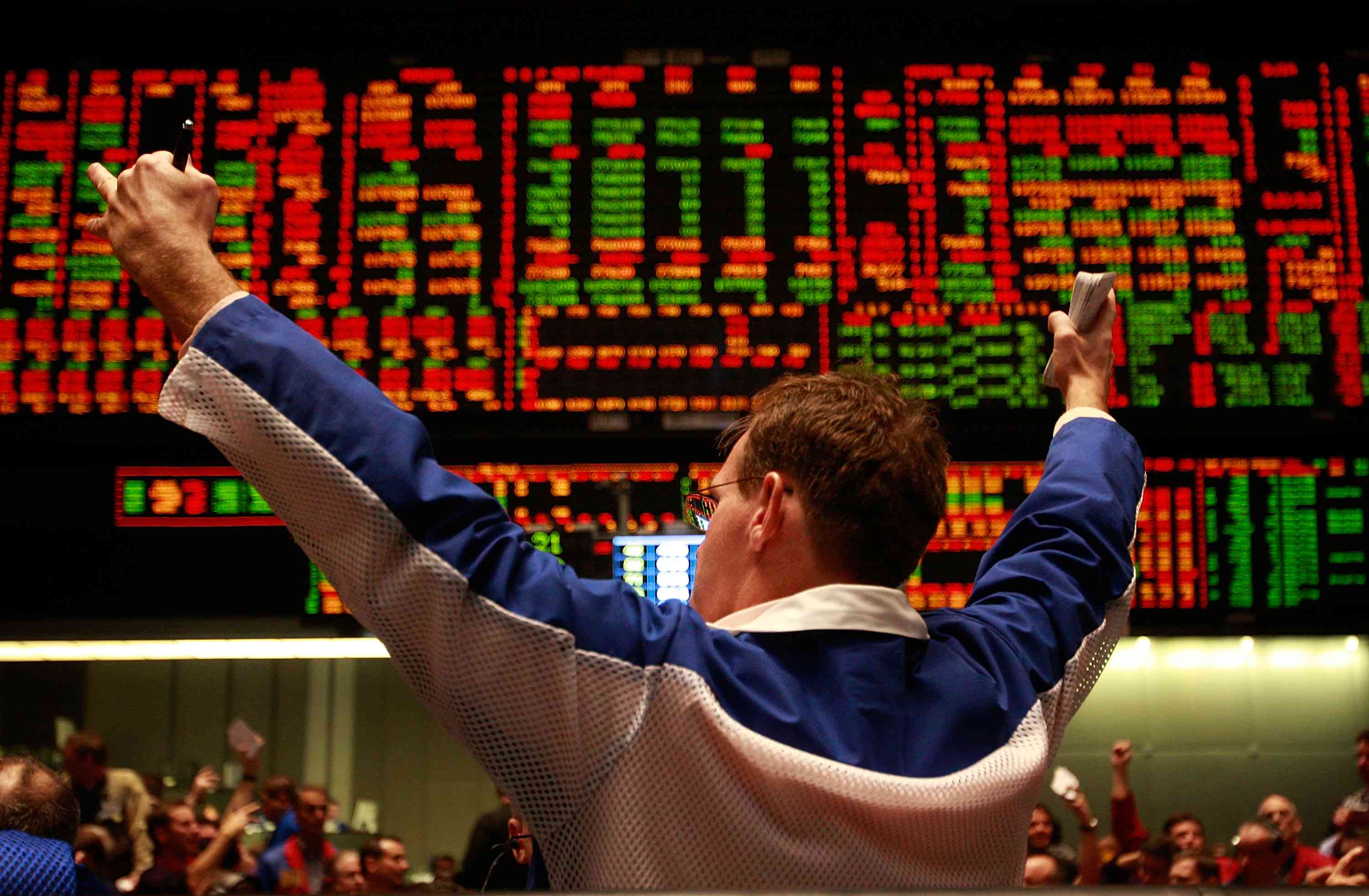 A male trader holds up both arms in front of a big screen displaying stock index information