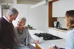 An older man and older woman stand in a kitchen with mugs talking to a younger women