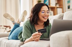 A woman lays on a couch, holding up a bank card and looking at a laptop.
