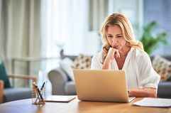 A woman in a white shirt sits at a desk at home with a laptop