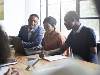 three colleagues in an office sitting at a table looking at a laptop and smiling