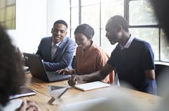 three colleagues in an office sitting at a table looking at a laptop and smiling