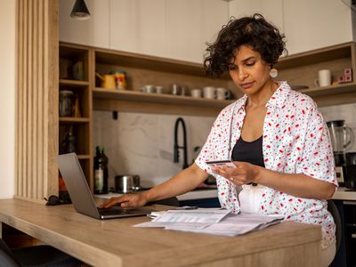 A woman stands in a kitchen at an island with a laptop and a credit card with documents sitting on the counter