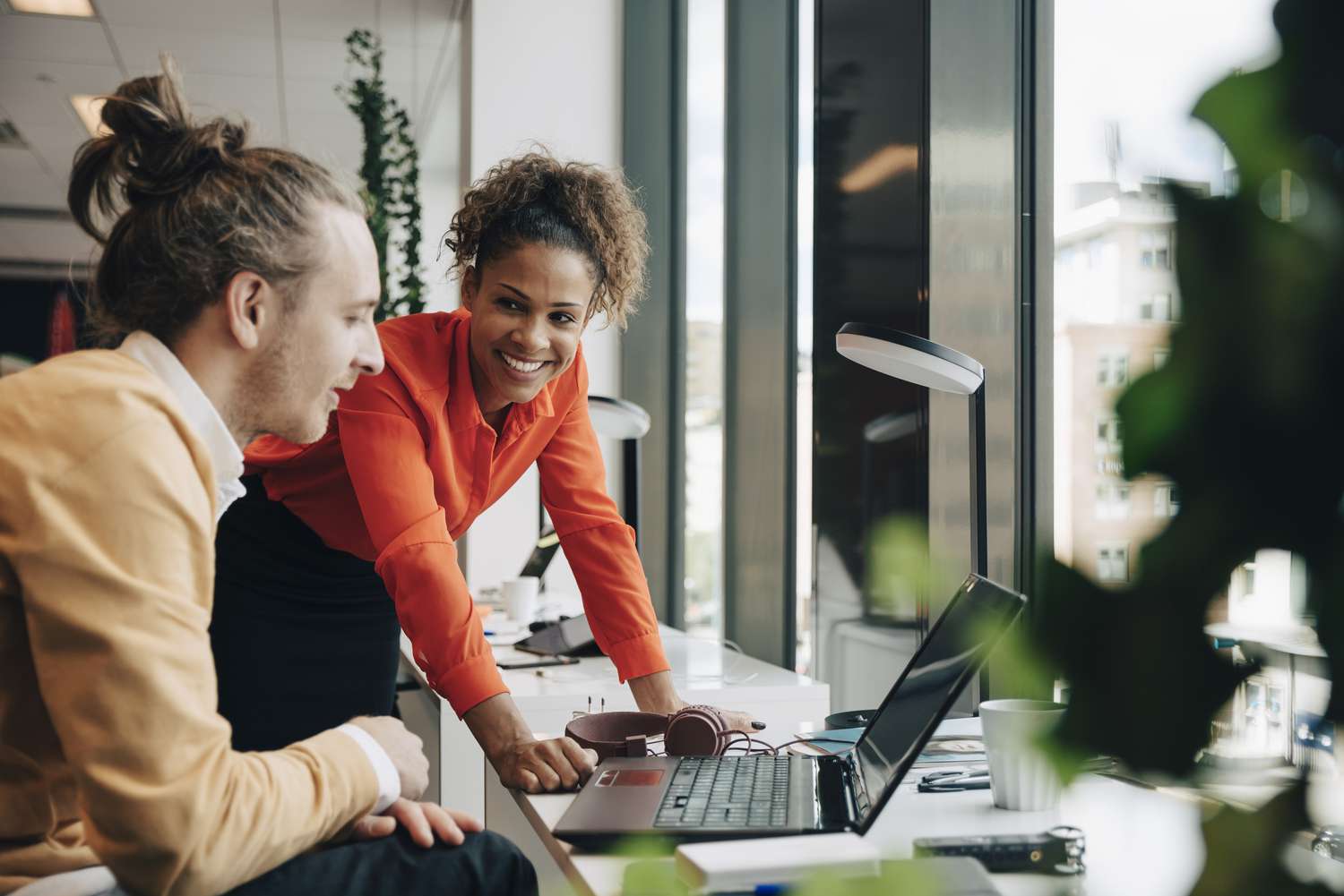 A man and a woman in an office laugh together over something they have seen on a laptop screen.