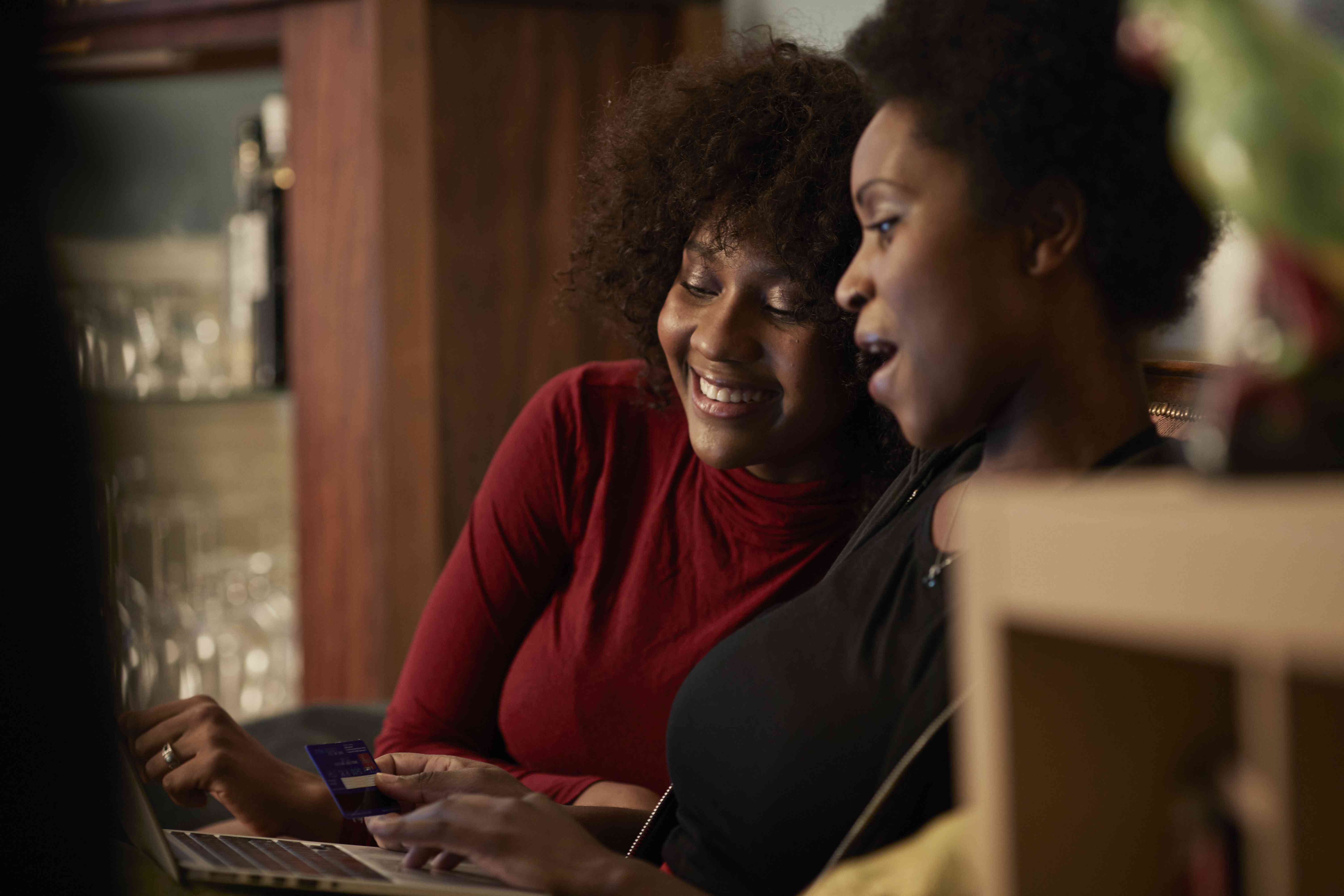 Two women sit on a couch with a credit card and a laptop