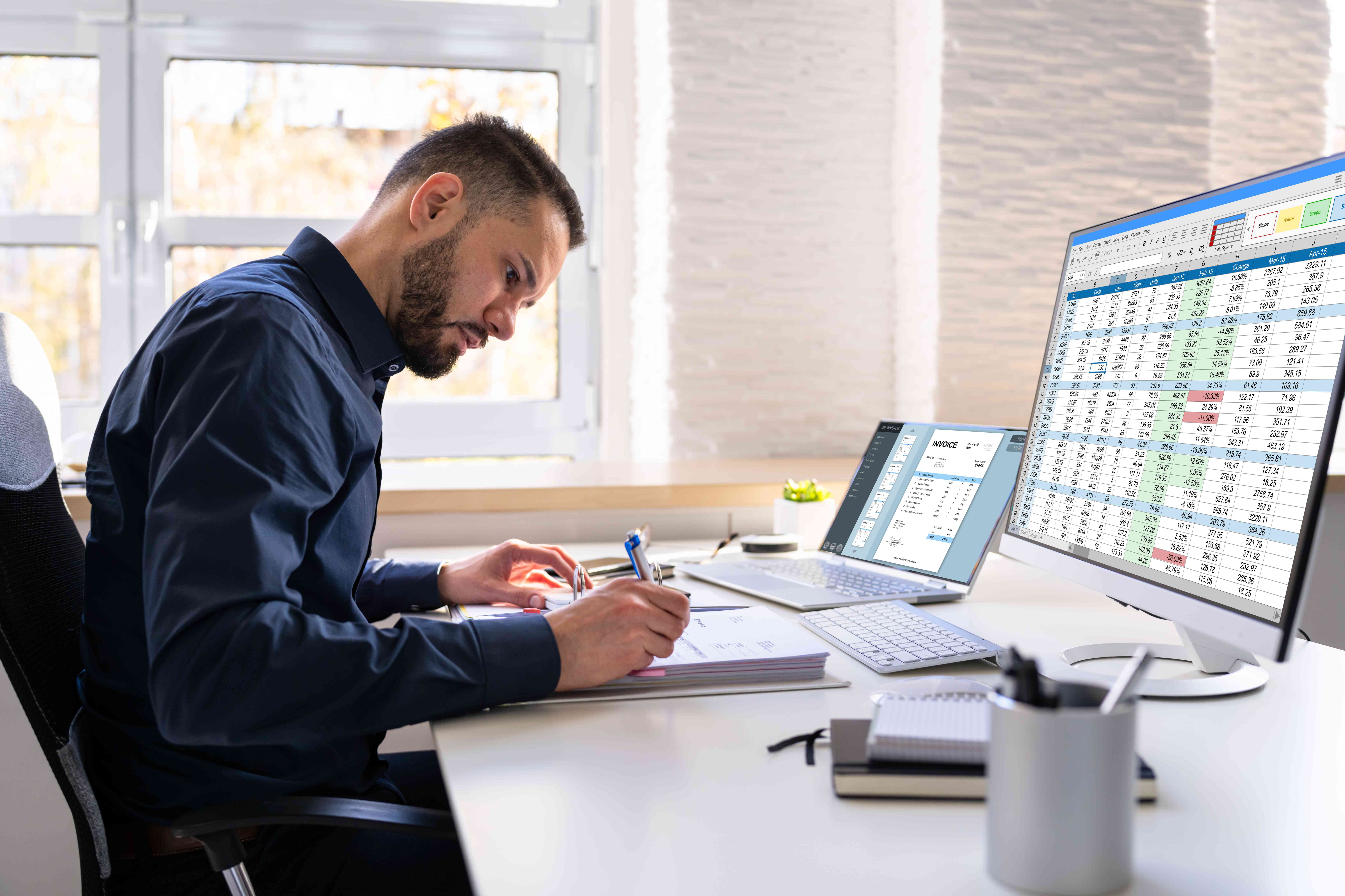 A man with a close-cropped beard seated at a desk writes on a notepad. Meanwhile a laptop screen and a desktop monitor display an invoice and a spreadsheet, respectively.