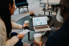 Two people sit on a sofa. They share a laptop, both looking at the statistics displayed, while they drink coffee.