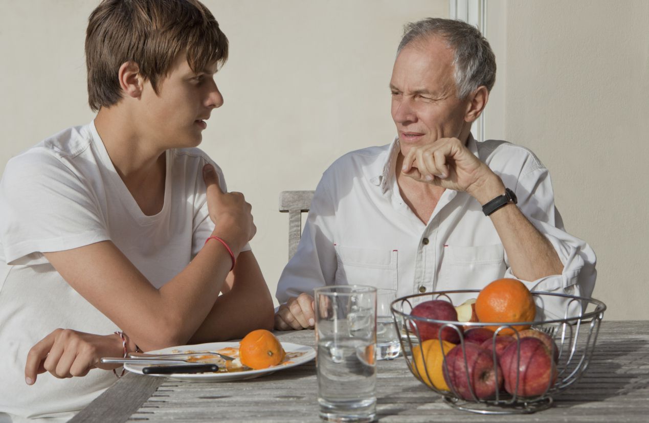 Older father talks with teen, sitting together at a table with a bowl of apples and oranges.