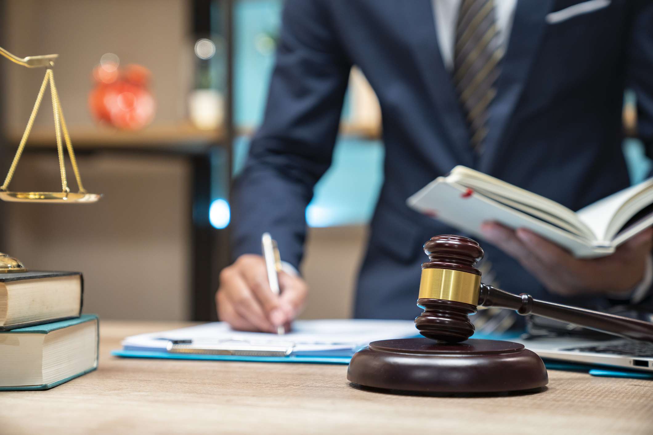 Cropped photo of a lawyer in a suit writing in a notepad with a scale, a gavel, and a block on the table.