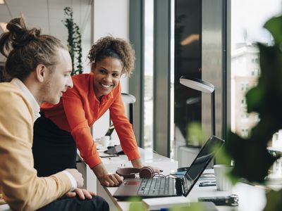 A man and a woman in an office laugh together over something they have seen on a laptop screen.