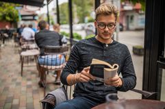 A man sits outside at a cafe at a table reading a book