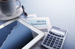 A digital tablet with a stock market chart on its screen rests on top of a 401(k) statement that sits on a desktop. A fountain pen, a calculator, and cup of coffee also frame the scene.