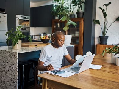 A man sits at a dining room table with a laptop, papers, and a calculator